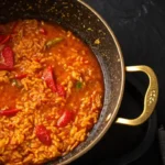 A close-up overhead image of a saffron-colored rice dish with red peppers in a black and gold pan.