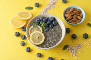 A bowl of chia seeds surrounded by lemon slices, blueberries, and almonds on a yellow background.