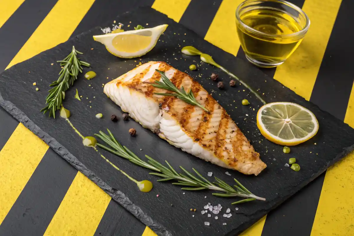 Grilled fish fillet served on a black slate plate with lemon wedges and rosemary.