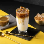 A tall glass of iced coffee with milk and coffee ice cubes sits on a black and yellow background.